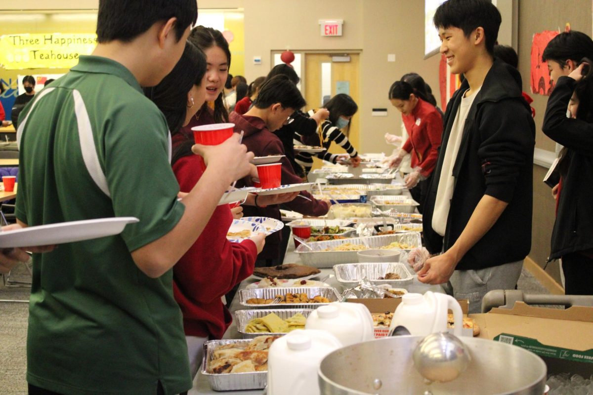 Joshua Lim ‘25, volunteering as a food server, smiles at the students in line to get food. Both traditional Chinese foods like egg and scallion crepes and cold noodles, and American foods like pizza are represented on the table. A large pot of milk tea sits at one end of the table and is ladled out into red cups by the volunteers.