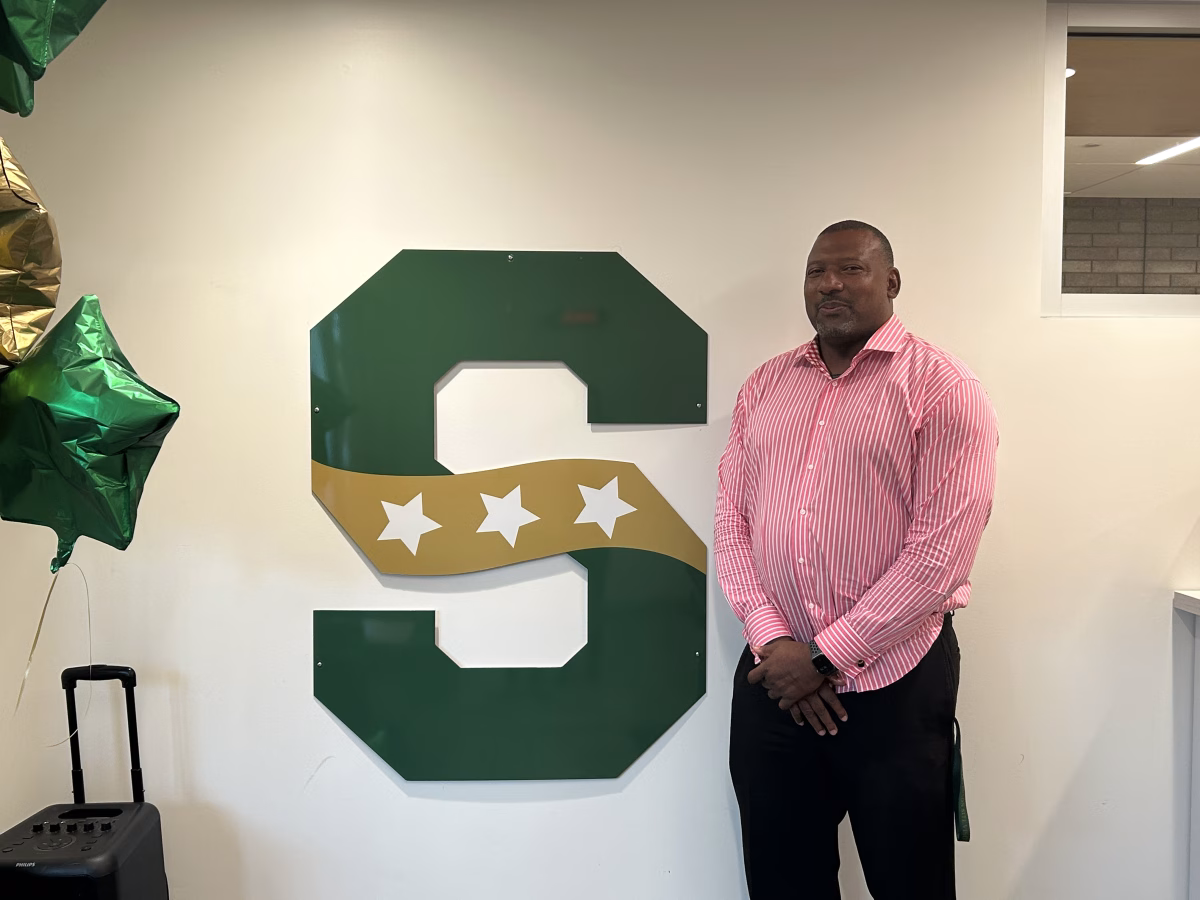 Athletic Director D’Wayne Bates stands in front of the SHS logo in the athletic office. Bates is happy to be working with the Stevenson community.